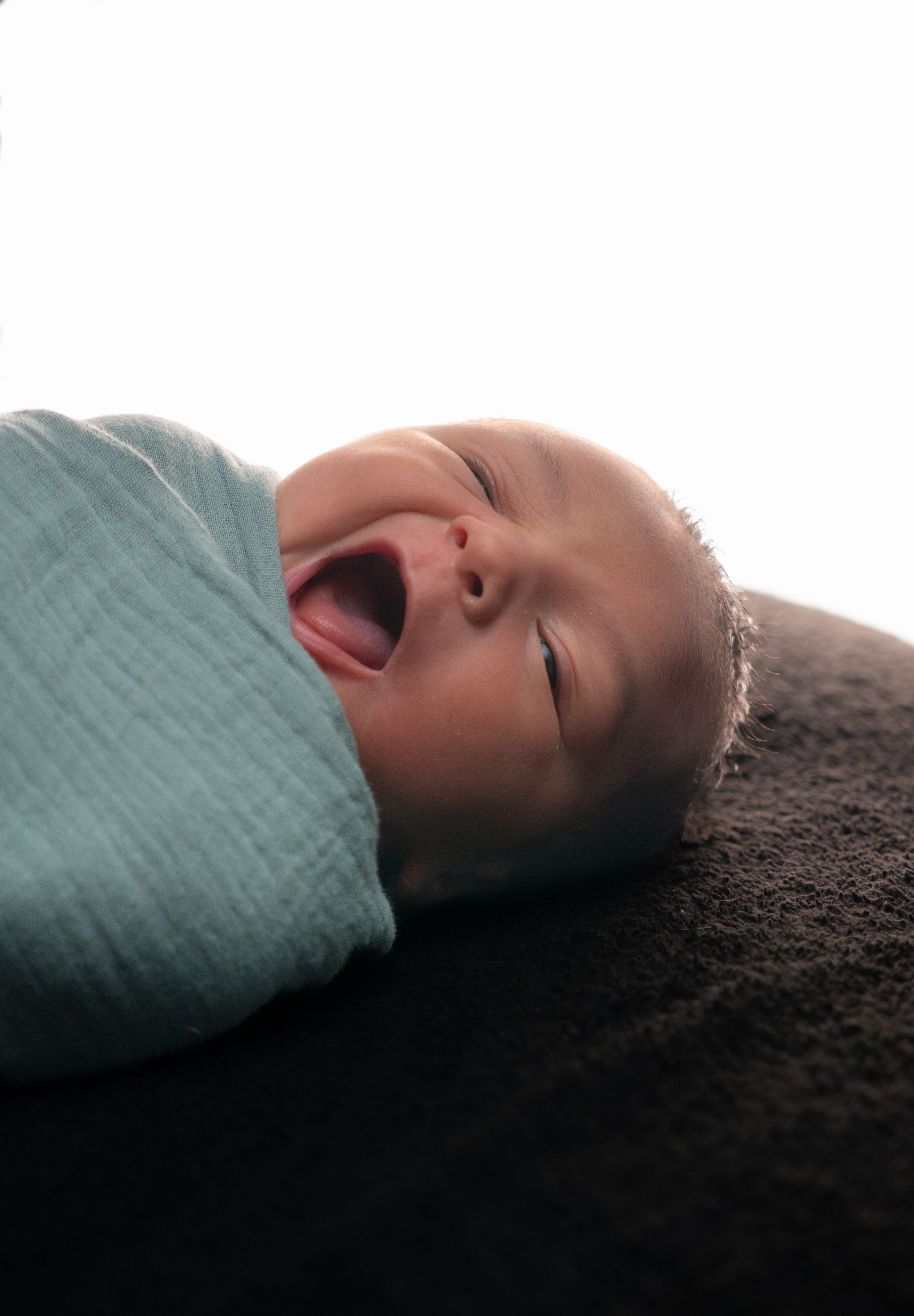 Swaddled newborn baby yawning with mouth wide open wrapped in a light blue muslin blanket on a dark surface.