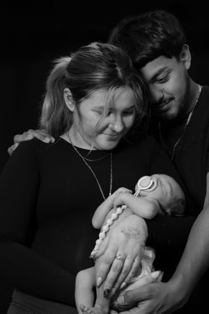New parents in black outfits tenderly hold their sleeping newborn baby together in a black and white portrait.