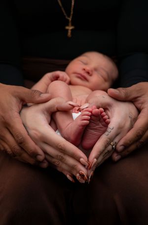 Parents form a heart shape with their hands around a smiling newborn baby's tiny feet in a warm-toned photo.