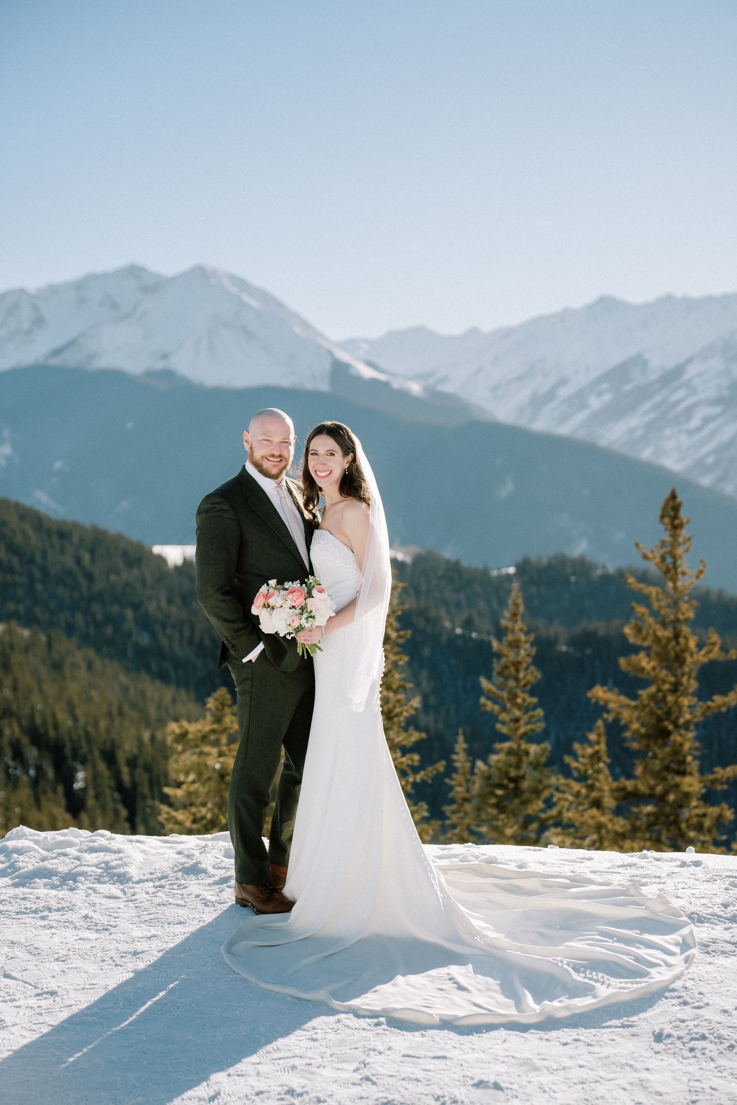 A couple in wedding attire stands together on a snowy mountain peak with evergreen trees and dramatic mountain views. Aspen elopement photo.