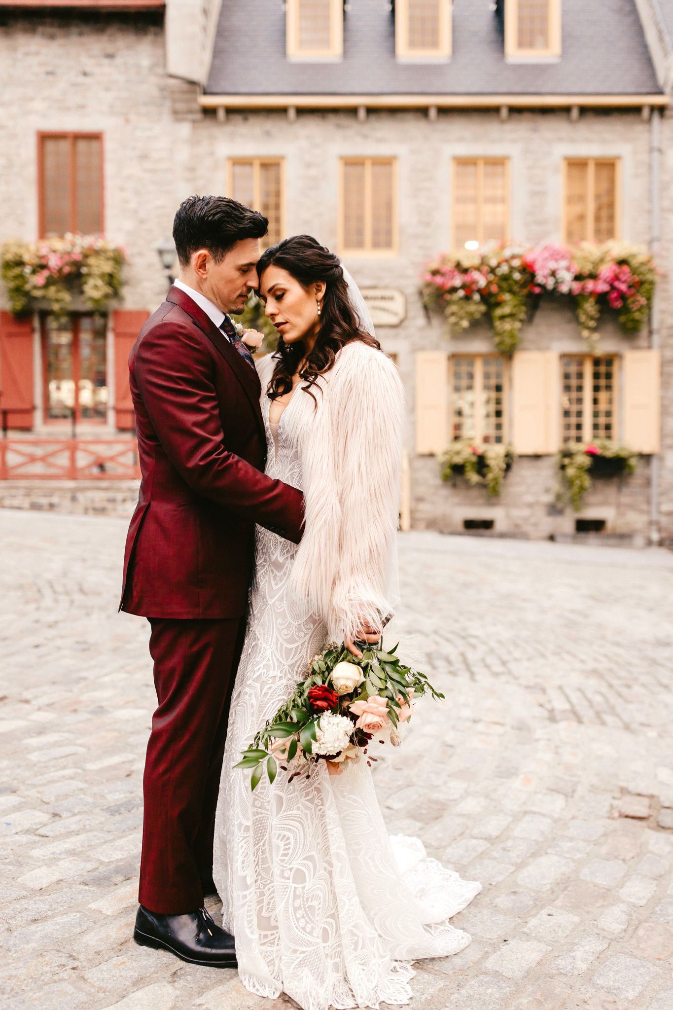 A couple in wedding attire embraces in front of a beautiful stone building with flower boxes.