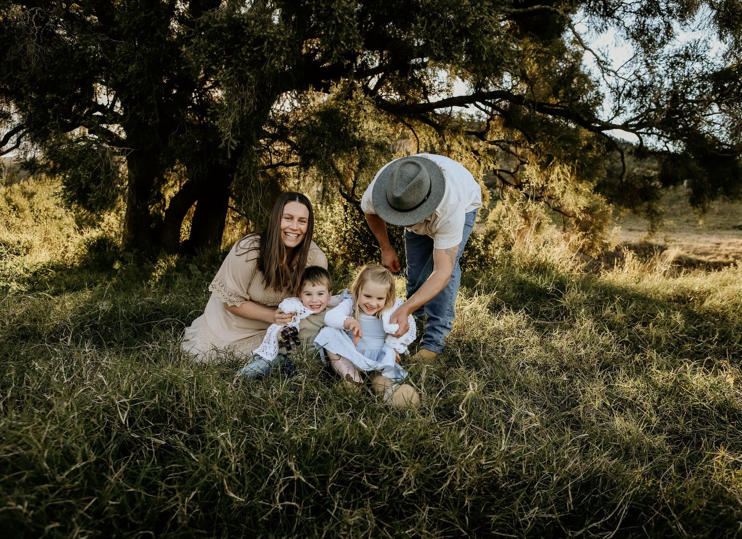 Family sits together on grassy hillside in golden sunlight during outdoor photo session.