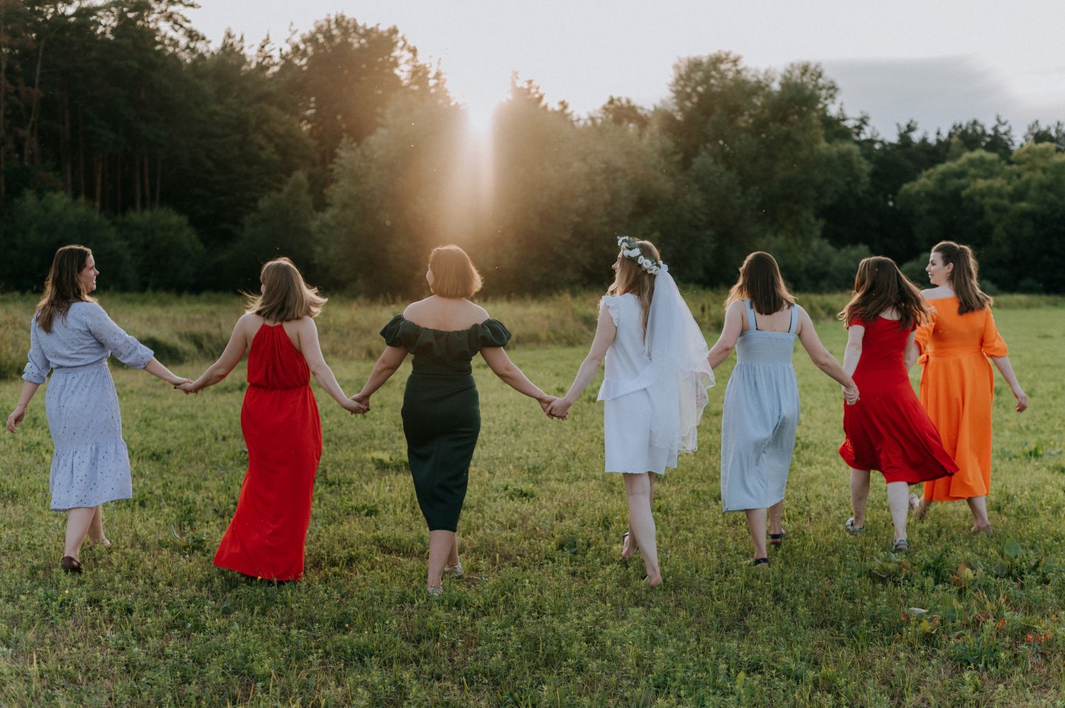 A group in colorful dresses joins hands while walking through a sunlit meadow together.