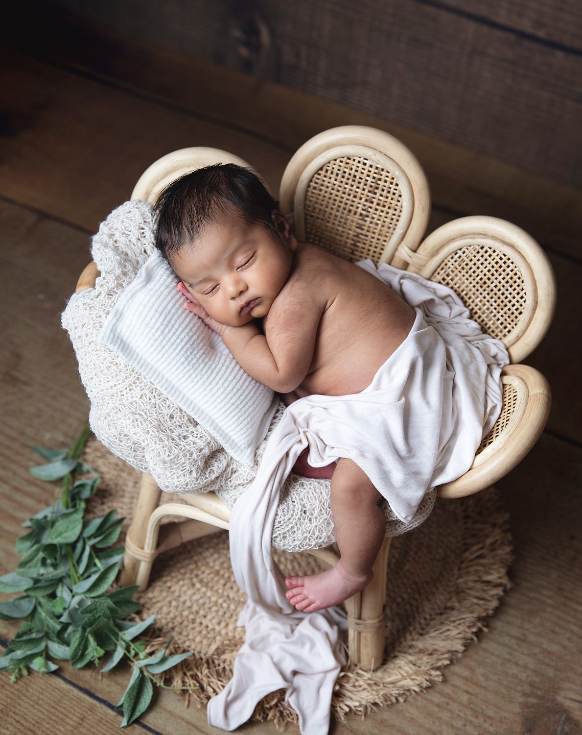 Peaceful sleeping newborn rests on decorative rattan chair with white pillow and lace accents against rustic wooden backdrop.