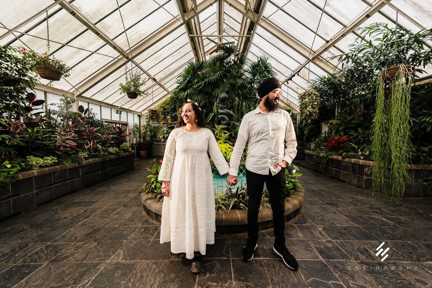 A couple holds hands while standing in a bright greenhouse filled with lush plants.