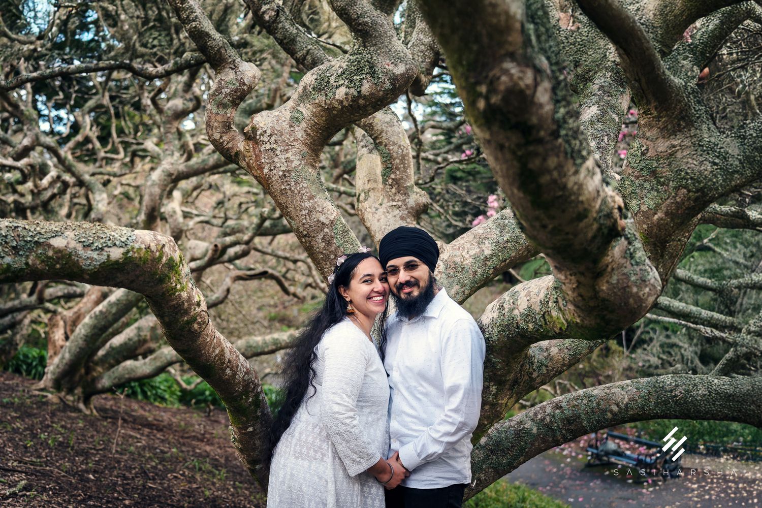 A couple poses together under the gnarled branches of an ancient-looking tree.