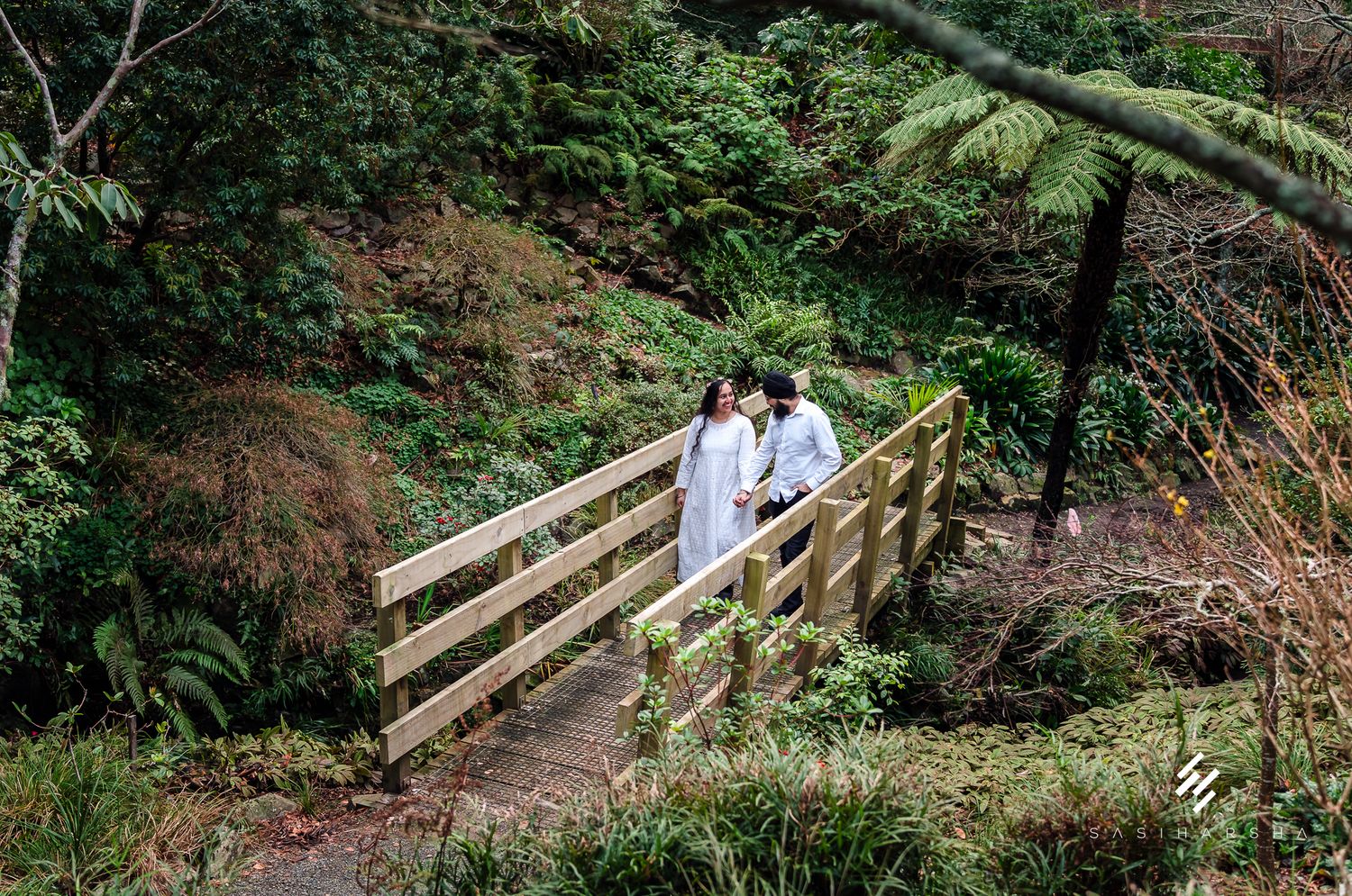 A couple stands on a wooden footbridge surrounded by lush forest greenery.
