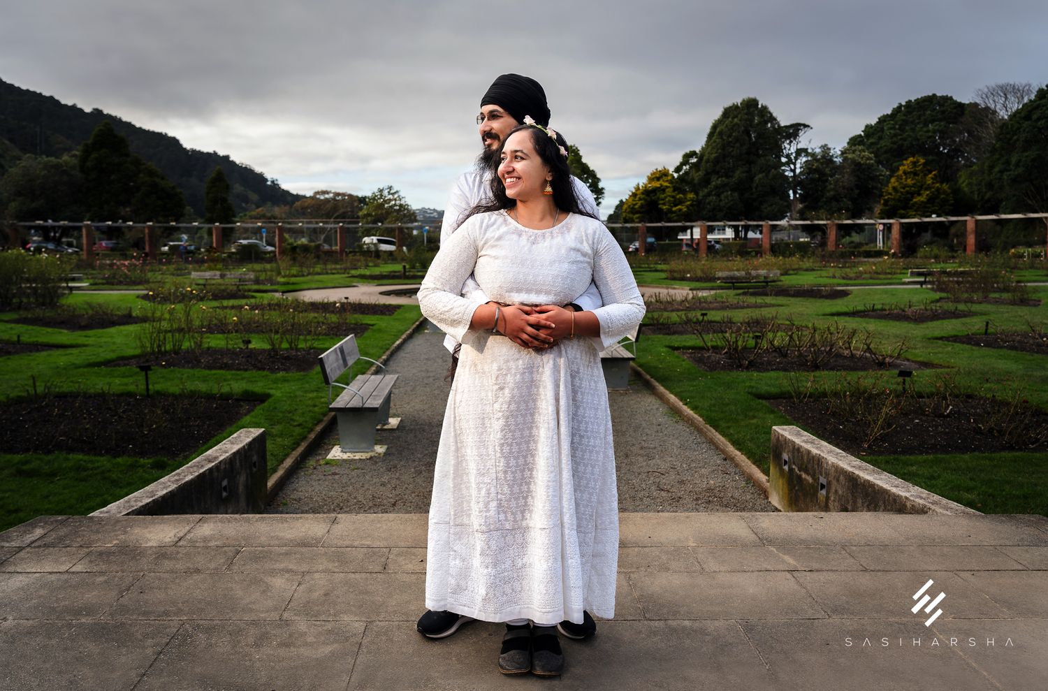 A couple embraces in a formal garden with cloudy skies overhead.