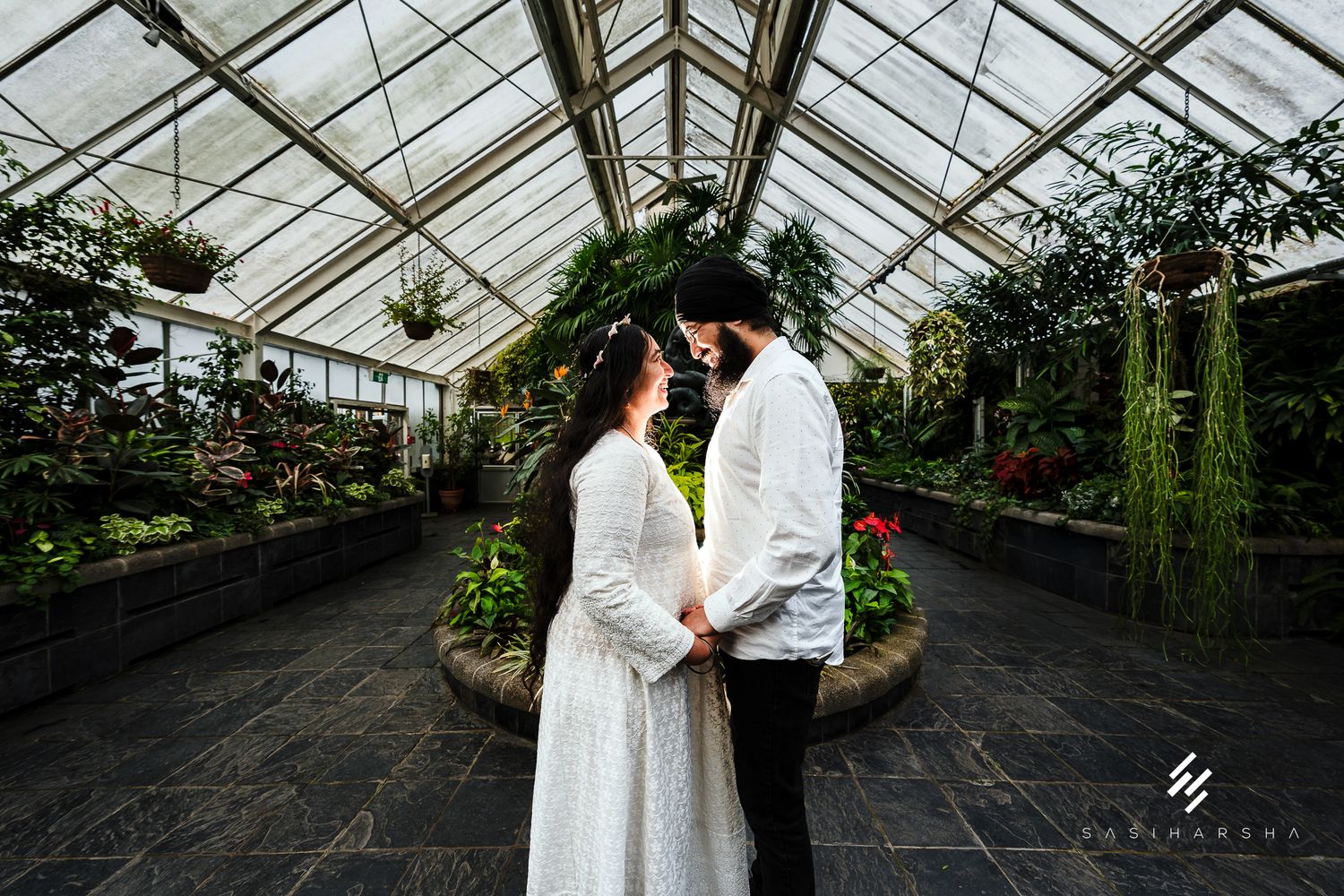 Interior of a large greenhouse with glass ceiling and potted plants lining the walkways.