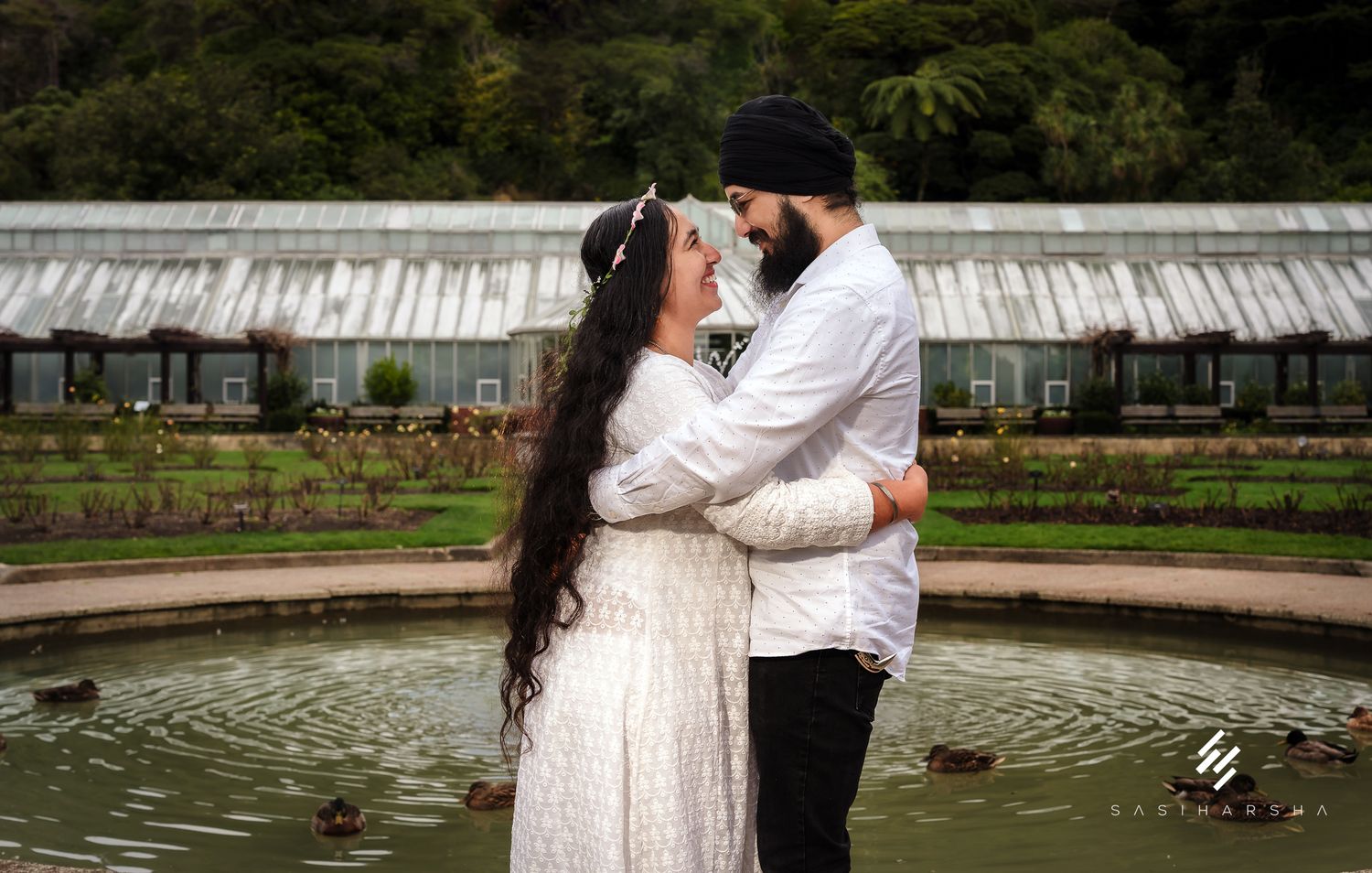 A couple embraces by a pond in a greenhouse garden with glass architecture in the background.