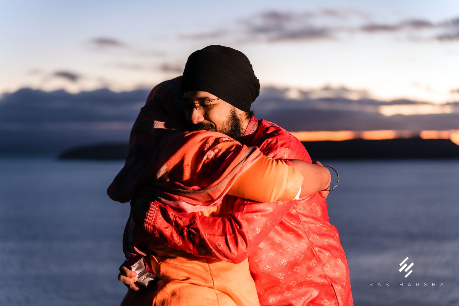 An intimate embrace in bright orange attire against a sunset seascape.