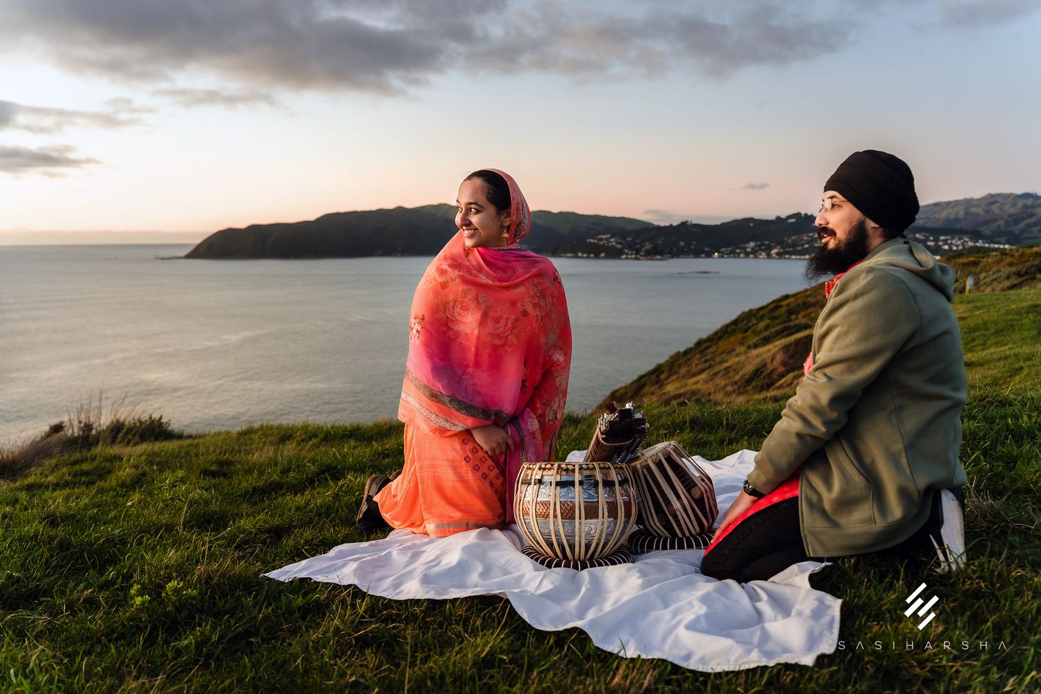 A couple sits with tabla drums on a blanket overlooking a coastal sunset.