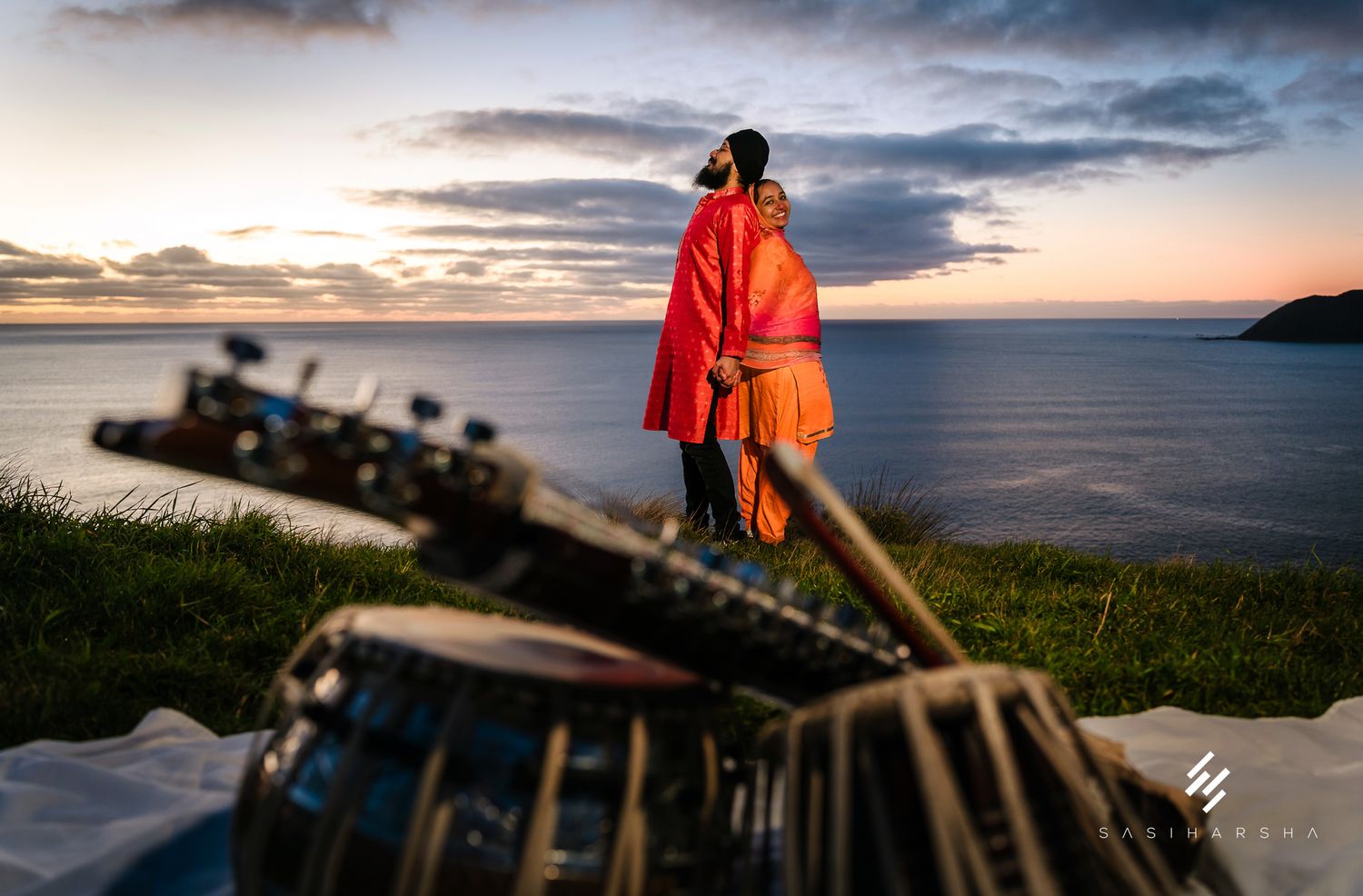 A silhouetted figure in orange attire stands against a dramatic sunset by the ocean with musical instruments.