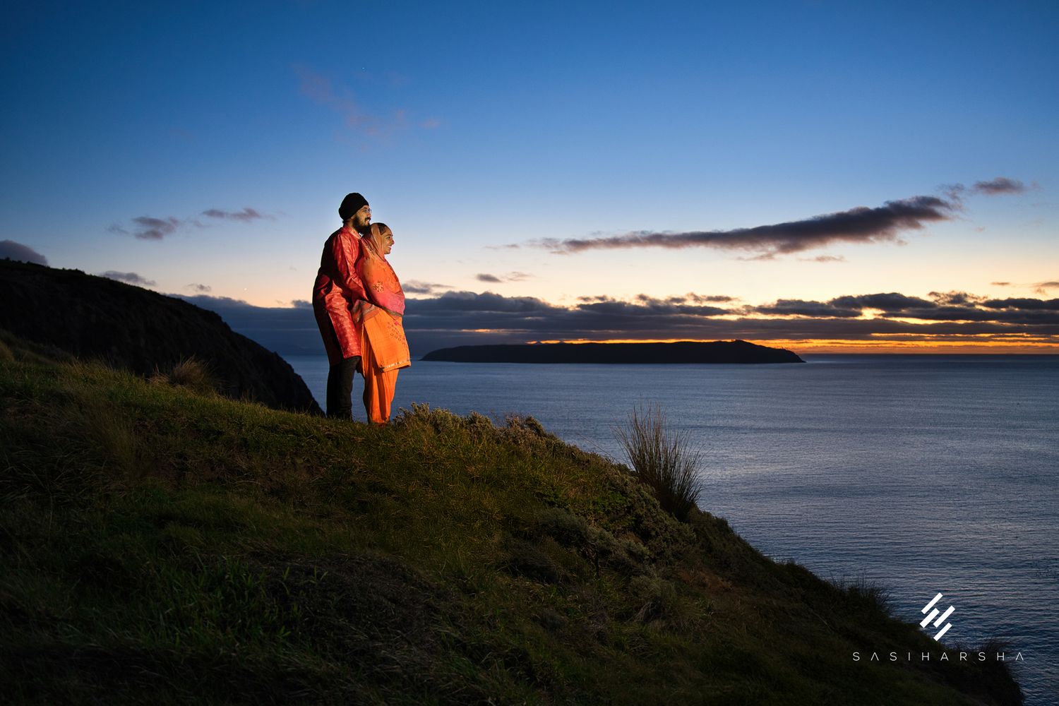Two figures in traditional attire stand on a cliff edge at sunset overlooking the ocean.