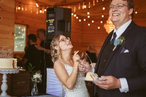 A wedding reception scene shows cake-sharing moment under warm string lights in a rustic wooden venue.