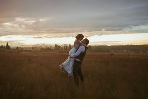 A couple embraces in a golden grass field as the sun sets behind dramatic clouds over a distant cityscape.