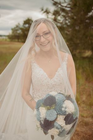 Bride wearing lace wedding dress and veil holds beautiful blue and white bouquet in outdoor setting.