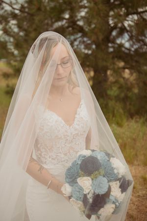 A bride in a lace gown and veil holds a blue and white bouquet during outdoor portraits.