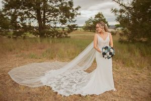 Elegant bride in a lace gown with cathedral veil poses in a rustic field holding a blue and white bouquet.