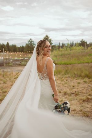 A bride in a lace wedding gown with a long flowing veil poses in an outdoor field at sunset.