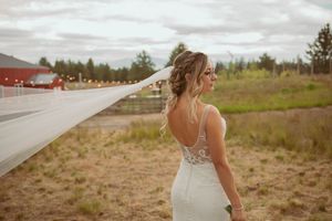 A bride in a lace wedding dress with flowing veil stands in a rustic outdoor field setting.