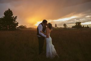 Silhouettes embrace in open field at sunset with dramatic orange sky and trees in background.