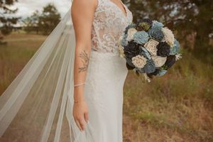 A detailed view of a wedding dress with lace bodice and flowing veil as someone holds a blue and white floral bouquet.