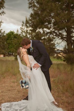 Romantic wedding photo of newlyweds kissing while holding a bouquet against a backdrop of pine trees.