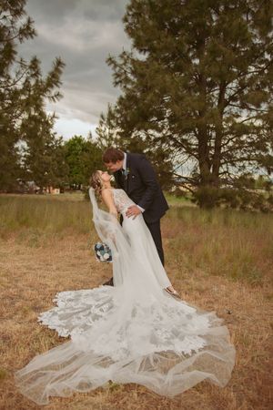 A romantic wedding couple embraces in a rustic outdoor setting with flowing white dress and veil spread on autumn grass.
