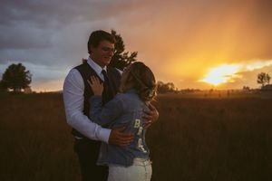 Two people embrace in formal attire while standing in a field during a dramatic sunset with dark clouds overhead.
