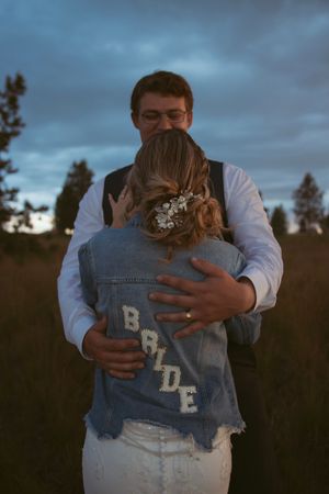 A couple embraces at dusk in a field, with the bride wearing a denim jacket with 'BRIDE' text.