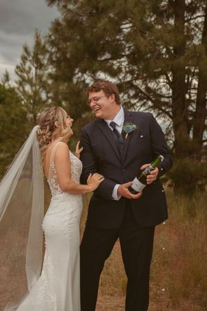 Wedding couple sharing champagne and laughter in an outdoor setting with pine trees.