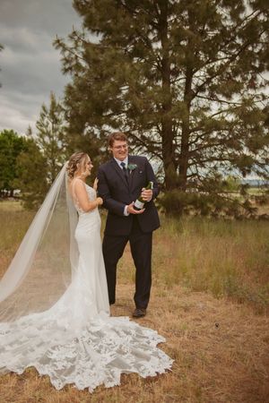 Couple celebrates during wedding photoshoot in rustic outdoor setting with tall pine trees and long train.