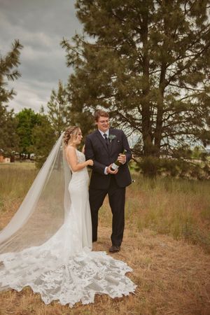 A bride in a long lace dress with flowing veil stands with groom in black suit outdoors among pine trees.