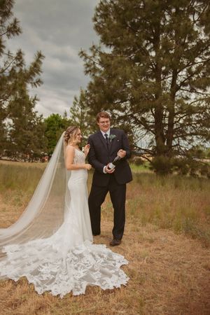 Newlyweds in elegant attire pose on grassy hillside with dramatic lace wedding dress train.
