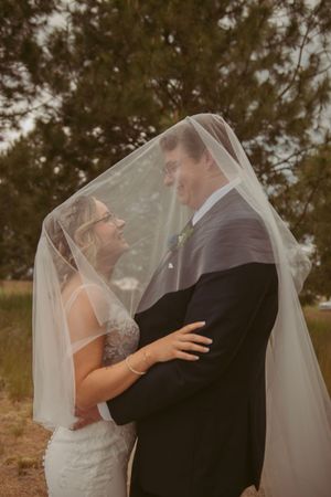Wedding couple shares intimate moment under flowing white veil against evergreen trees and cloudy sky.