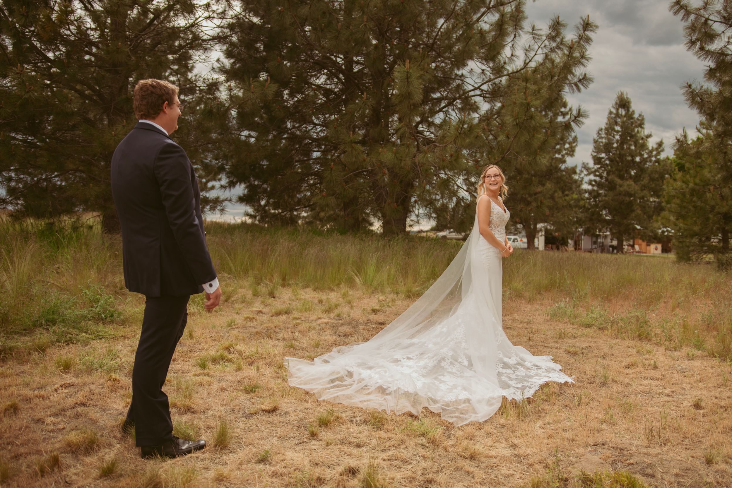 Wedding couple poses outdoors in a grassy field with pine trees while the bride's elegant white dress trails behind her.