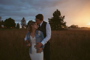 A couple shares an intimate moment in a field at sunset with soft golden light streaming through dramatic clouds.