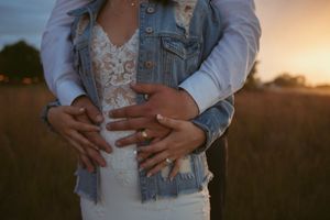 A pair stands close together at sunset wearing a white lace dress and denim jacket with rings visible on their hands.