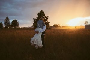 A silhouetted couple embraces in a sunset-lit field with tall grass and towering evergreen trees framing the scene.
