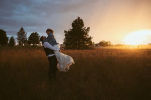 Couple embraces in a golden sunset field with tall grass and evergreen trees in the background.