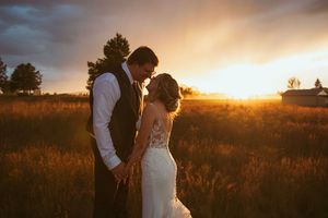 A romantic wedding couple embraces in a golden sunset field with tall grass and trees silhouetted against the sky.