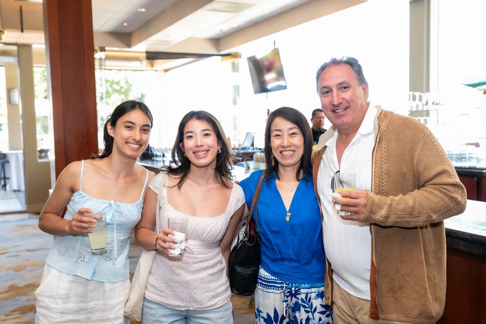A group of friends gathered at a bar or restaurant counter, smiling and holding up drinks in a celebratory toast.