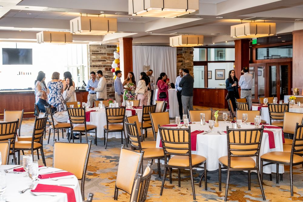 A group gathers in a bright dining room with wooden furniture and pendant lights during a social event.