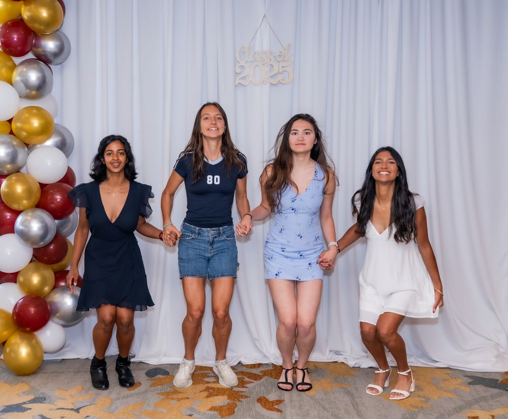 Four friends in casual summer outfits sitting in a row against a white backdrop with festive gold and maroon balloons.