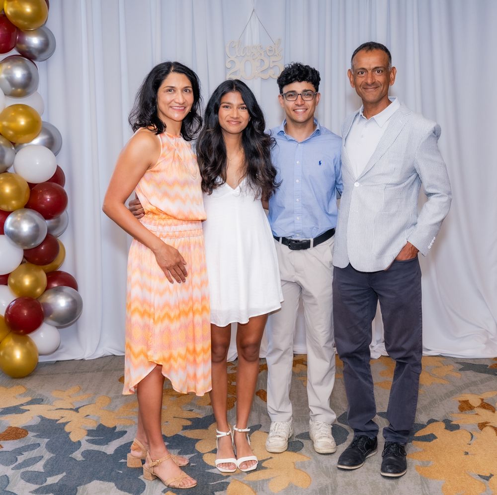 A family poses together in formal attire against a white backdrop with festive balloon decorations.