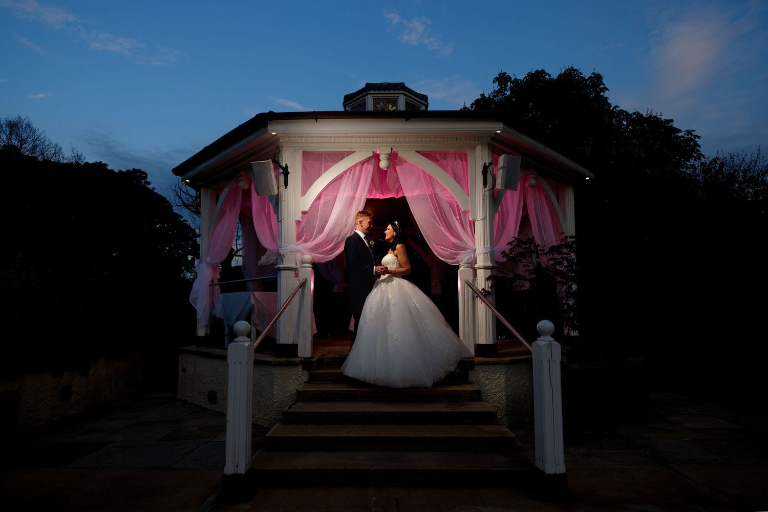 A couple shares a romantic dance under a gazebo decorated with pink curtains at dusk.