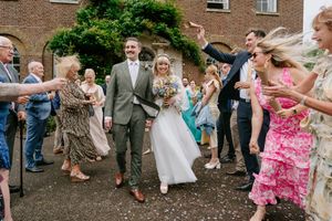 Wedding guests in elegant attire dance and celebrate during an outdoor reception near a brick building.