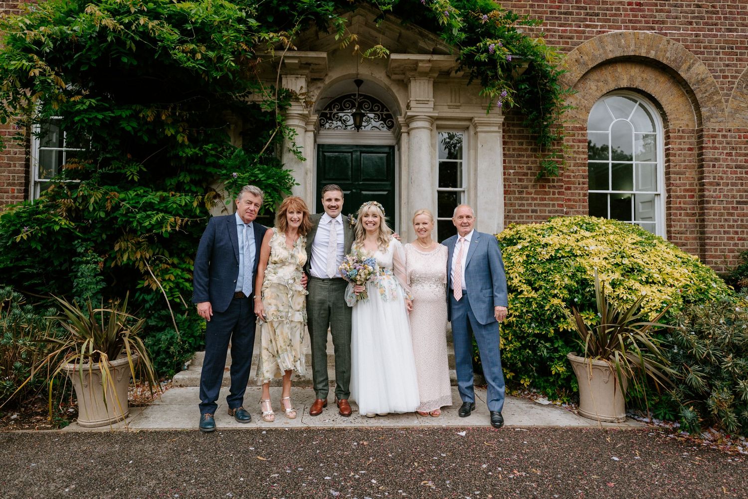 Wedding party stands together at romantic brick venue entrance with greenery and arched doorway.