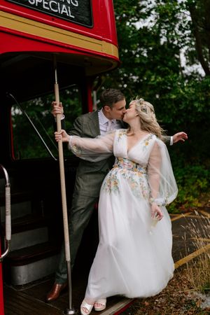 A couple in wedding attire poses with a classic red double decker bus in the background.